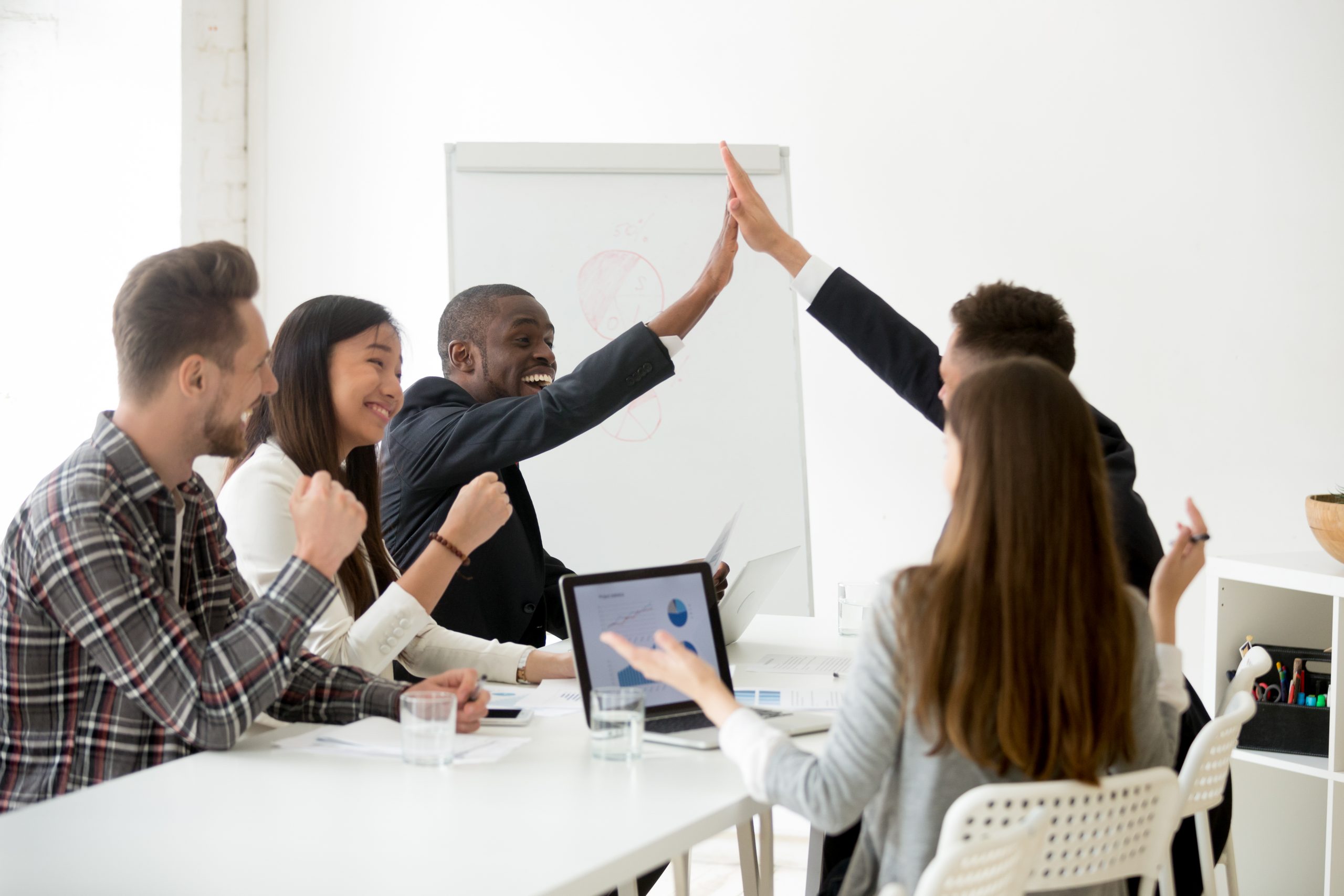 Excited diverse executive colleagues or partners giving high five at team meeting, happy smiling multiracial businessmen celebrating good teamwork result motivated by great achievement or victory at a business meeting.